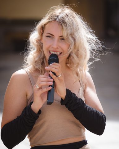 Junge Frau mit Locken hält ein Mikrofon und spricht lächelnd in die Kamera.